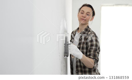 Woman wearing protective gloves and checkered shirt is spreading finishing putty across white wall surface, smoothing damaged areas before painting interior renovation work Woman wearing protective gloves and checkered shirt is spreading finishing putty across white wall surface, smoothing damaged areas before painting interior renovation work 131868559