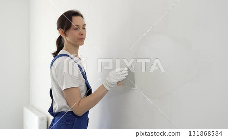 Female construction worker spreading finishing putty on white wall, wearing protective gloves and blue work overall while smoothing surface with putty knife for precise home repair 131868584