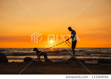 Man standing on rocks at the beach holding a leash with a German Boxer dog, both watching the sunset over the ocean. Man standing on rocks at the beach holding a leash with a German Boxer dog, both watching the sunset over the ocean. 131869008
