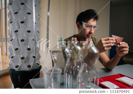 Agricultural technician wearing safety glasses examining fertilizer packet while preparing to nourish plants in vertical hydroponic tower, surrounded by measuring equipment, recycled plastic bottles. 131870491