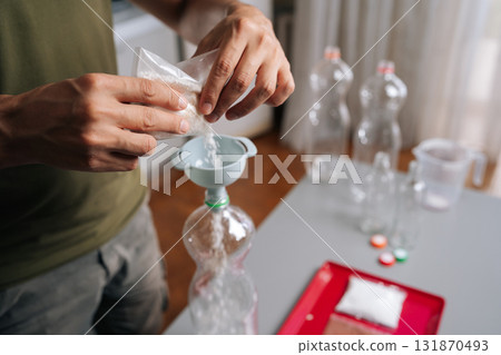 Close-up hands of unrecognizable gardener pouring organic fertilizer into plastic bottle using funnel, preparing nutrients for plants in home kitchen setting, demonstrating diy approach to gardening 131870493