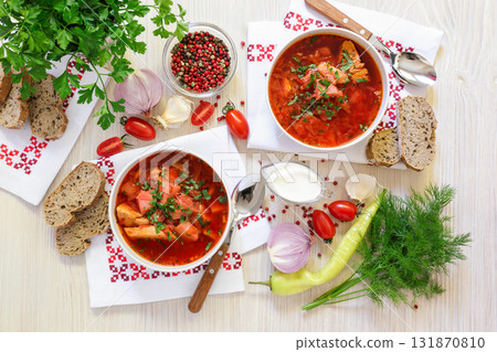 Two bowls of Ukrainian borscht on embroidered napkins, bread, sour cream, spices, onions, garlic, tomatoes, parsley, dill on a white wooden background. Top view 131870810