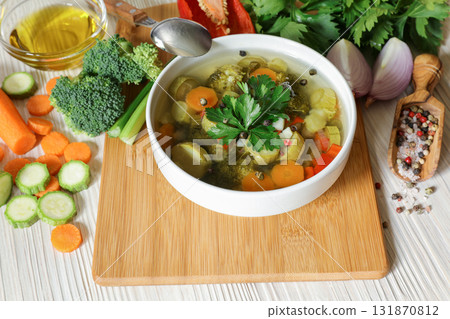 Light vegetable soup with ingredients of zucchini, broccoli, carrot, celery, pepper, onion, spices and a little olive oil on a white wooden background. 131870812