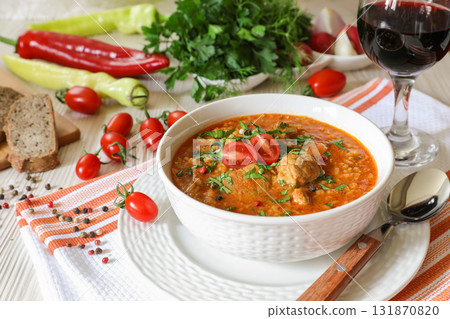 Traditional Georgian soup Kharcho with meat and rice in a white bowl next to bread, tomatoes, peppers, herbs on a white orange tablecloth. Closeup traditional table setting. Traditional Georgian soup Kharcho with meat and rice in a white bowl next to bread, tomatoes, peppers, herbs on a white orange tablecloth. Closeup traditional table setting. 131870820