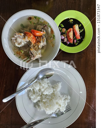 This vibrant overhead photograph beautifully captures an Indonesian meal featuring a generous bowl of clear, savory lamb or goat rib soup (Sop Iga Kambing or Sop Kambing) garnished with sliced tomatoe 131873347