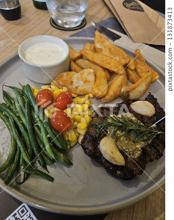 A delicious grilled steak is beautifully plated with roasted garlic, a sprig of rosemary, a side of golden potato wedges, steamed green beans, sweet corn, and bright cherry tomatoes, 131873413