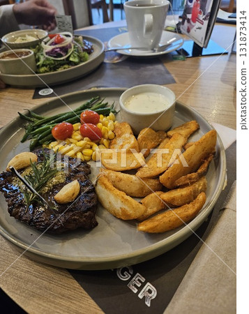 A delicious grilled steak is beautifully plated with roasted garlic, a sprig of rosemary, a side of golden potato wedges, steamed green beans, sweet corn, and bright cherry tomatoes, 131873414