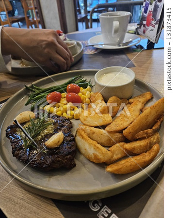 A delicious grilled steak is beautifully plated with roasted garlic, a sprig of rosemary, a side of golden potato wedges, steamed green beans, sweet corn, and bright cherry tomatoes, 131873415