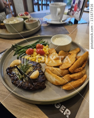 A delicious grilled steak is beautifully plated with roasted garlic, a sprig of rosemary, a side of golden potato wedges, steamed green beans, sweet corn, and bright cherry tomatoes, 131873416