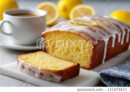 Homemade lemon drizzle cake with icing. Classic lemon pound cake with citrus glaze and zest. Whole loaf, side view, close up. Fresh homemade baked dessert with lemon slices. Blurred tea set background 131874613