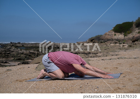 Elderly man practicing yoga by the ocean shore, resting in Balasana. Elderly man practicing yoga by the ocean shore, resting in Balasana. 131876310