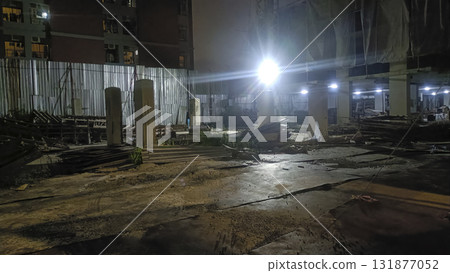 Construction site at night illuminated by bright floodlight and scattered building materials, concrete columns and temporary fencing Construction site at night illuminated by bright floodlight and scattered building materials, concrete columns and temporary fencing 131877052