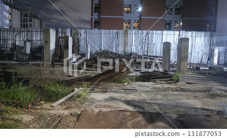 Abandoned construction site at night with corrugated metal fence, concrete pillars, scattered rebar and debris under moody lighting 131877053