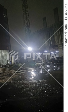 Dimly lit construction site at night with wet reflective ground and bright floodlight creating moody atmosphere Dimly lit construction site at night with wet reflective ground and bright floodlight creating moody atmosphere 131877054