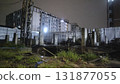 Dilapidated construction site with concrete pillars and overgrown grass under night sky, moody urban scene with apartment building lights 131877055