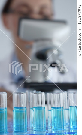 Close up of test tubes with blue liquid while scientist focusing on analyzing samples under a microscope in a laboratory setting. Medicine, healthcare and science concept 131877072