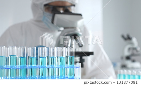 Medical scientist wearing protective suit using microscope while analyzing samples, conducting research in laboratory environment. Close up of test tubes with blue liquid 131877081