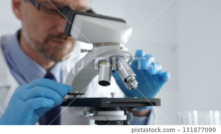 Close up of male scientist in white coat and blue gloves adjusting microscope, conducting pharmaceutical research in modern laboratory. Medicine and science concept Close up of male scientist in white coat and blue gloves adjusting microscope, conducting pharmaceutical research in modern laboratory. Medicine and science concept 131877099
