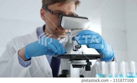 Male scientist in white coat and blue gloves adjusting microscope, conducting pharmaceutical research in modern laboratory. Medicine, healthcare and science concept Male scientist in white coat and blue gloves adjusting microscope, conducting pharmaceutical research in modern laboratory. Medicine, healthcare and science concept 131877100