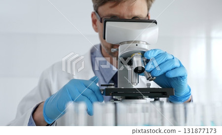 Male scientist in white coat and blue gloves adjusting microscope, conducting pharmaceutical research in modern laboratory. Medicine, healthcare and science concept 131877173