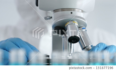 Scientific researcher or virologist wearing protective gear is tuning microscope lenses opposite the lab tubes with blue liquid in laboratory setting. Medicine, healthcare and science concept 131877196
