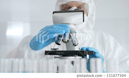 Scientific researcher wearing protective gear adjusting microscope, analyzing lab samples with precision in sterile research environment. Medicine, healthcare and science concept 131877304