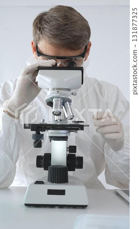 A male medical virologist in a medical gown and gloves examines viral samples under a microscope while conducting research in a sterile laboratory setting A male medical virologist in a medical gown and gloves examines viral samples under a microscope while conducting research in a sterile laboratory setting 131877325