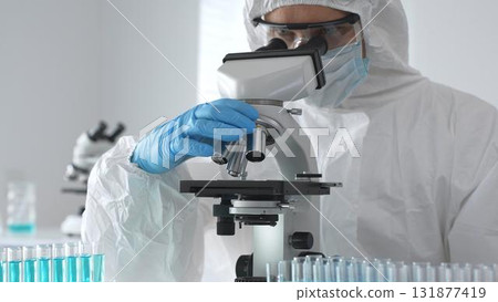 Medical virologist in protective hazmat gear examining viral samples under a microscope while conducting research in a sterile laboratory environment 131877419