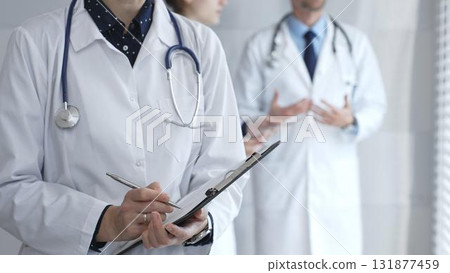 A medical professional with a stethoscope writes on a clipboard while a doctor and female assistant collaborate in a clinic, discussing patient data and healthcare information 131877459