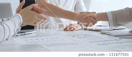 Business partners shaking hands at modern office desk with financial reports and laptop, colleagues clapping hands celebrating successful teamwork and agreement 131877501