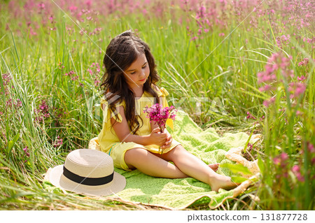 Pretty girl in a yellow dress, sits on a green knitted plaid in blossom field Pretty girl in a yellow dress, sits on a green knitted plaid in blossom field 131877728