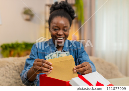 African American young woman opens birthday gift box with envelope of cash dollars sitting at home African American young woman opens birthday gift box with envelope of cash dollars sitting at home 131877909