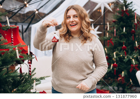 Happy woman holding miniature house toy. Concept of real estate, home ownership, and housing market. Young female decorating Christmas tree at home and smiling. Winter time and holidays 131877960