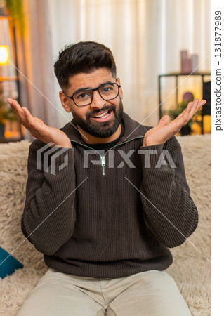 Indian man at sofa spreads hands in confusion and surprise at home, asking what with puzzled face Indian man at sofa spreads hands in confusion and surprise at home, asking what with puzzled face 131877989