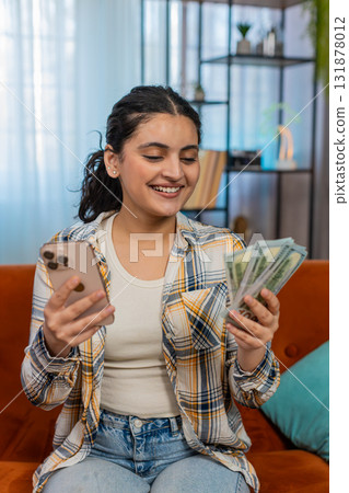 Indian woman on sofa with smartphone counting cash dollars planning expenses online transfers Indian woman on sofa with smartphone counting cash dollars planning expenses online transfers 131878012