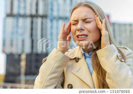 Sick mature Caucasian businesswoman feels headache while standing against buildings on city street 131878088