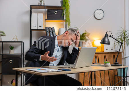Exhausted young businessman in formal suit working on laptop computer sitting at desk in home office 131878143