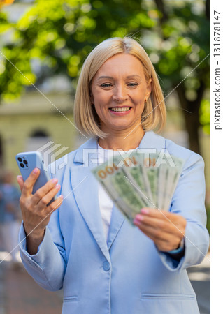 Caucasian mature businesswoman on city street showing earnings cash money with smartphone in hands 131878147