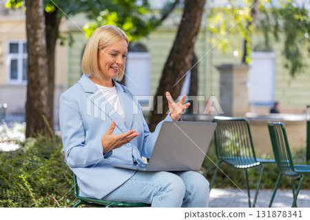 Mature businesswoman talking on video call using laptop while sitting on bench in city street Mature businesswoman talking on video call using laptop while sitting on bench in city street 131878341