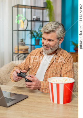 Mature man sitting at table with laptop and gamepad, playing video games and eating popcorn at home Mature man sitting at table with laptop and gamepad, playing video games and eating popcorn at home 131878402