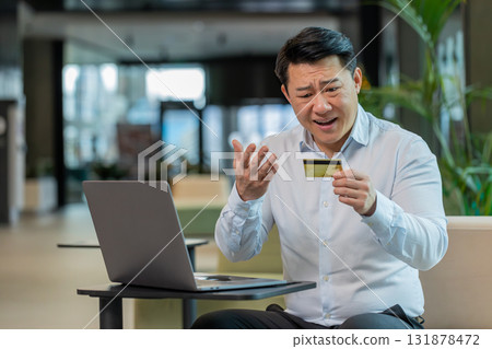 Angry Chinese mature businessman holding credit bank card sitting at modern workplace in office 131878472