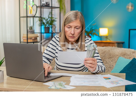 Mature woman planning family budget on laptop reviewing documents, counting money on table at home Mature woman planning family budget on laptop reviewing documents, counting money on table at home 131878475