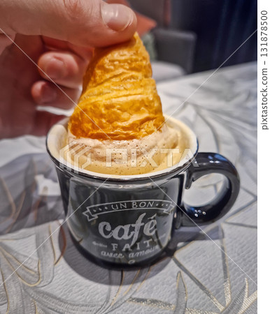 Close-up person hand dipping a mini croissant into a black coffee mug filled with frothy, caramel colored espresso drink. Good beverage with rich crema for a morning in french style 131878500