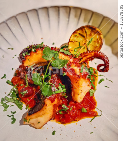 Close-up rich gourmet seafood dish. Tender charred octopus tentacles on a bed of chunky tomato sauce, garnished with fresh cilantro or parsley and caramelized lemon slice on a large, white plate Close-up rich gourmet seafood dish. Tender charred octopus tentacles on a bed of chunky tomato sauce, garnished with fresh cilantro or parsley and caramelized lemon slice on a large, white plate 131878505