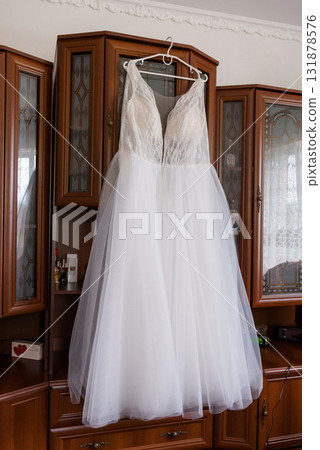 Elegant White Wedding Dress Hanging Before Wooden Cabinet. Bridal Gown, Tulle Skirt, Romantic Atmosphere, Interior Shot, Natural Light Elegant White Wedding Dress Hanging Before Wooden Cabinet. Bridal Gown, Tulle Skirt, Romantic Atmosphere, Interior Shot, Natural Light 131878576