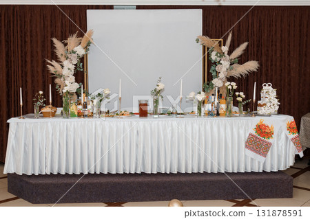 Elegant Wedding Table Setup: White Floral Arrangement, Pampas Grass, Gold Accents, Romantic Decor, and Traditional Ukrainian Textile Backdrop 131878591