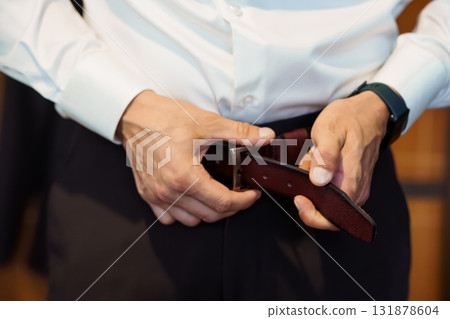 Man Fastening Brown Leather Belt with Silver Buckle Over Dark Pants, Formal Attire, Groom Preparing, Close-Up, Rich Texture, Studio Shot 131878604