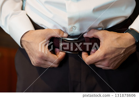 Man Fastening Dark Brown Leather Belt with Silver Buckle over Gray Trousers, Business Attire, Close-Up, Formal Style, Focus on Hands 131878606