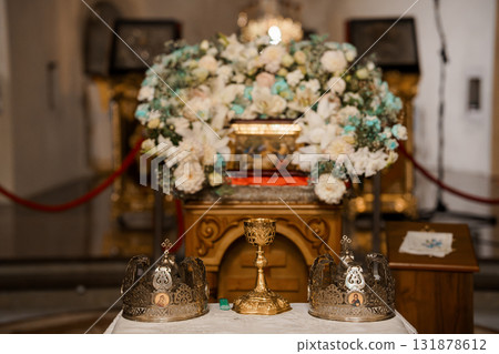 Orthodox Wedding Ceremony Altar with Crowns and Flowers: Golden Chalice, White Floral Arrangement, Religious Icons, Red Accents, and Elegant Chapel Interior. 131878612