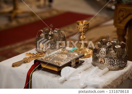 Orthodox Christian Coronation Ritual: Golden Crown, Holy Book, Chalice and Cross, Liturgical Objects on White Cloth, Deep Red Rug, Sacred Church Interior. Orthodox Christian Coronation Ritual: Golden Crown, Holy Book, Chalice and Cross, Liturgical Objects on White Cloth, Deep Red Rug, Sacred Church Interior. 131878618